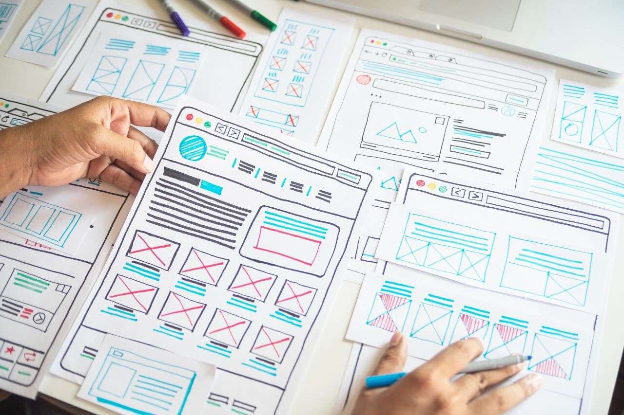 UI/UX-designer jobber med mobilapp-design ved skrivebord – omgitt av skisser og kaffekopp.